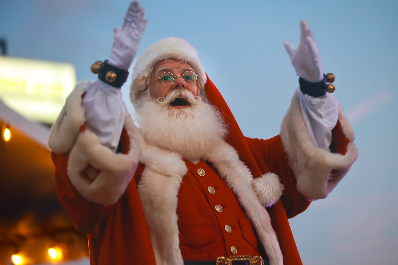 Santa Claus with both gloved hands raised, calling out from the top of a fire truck at dusk.