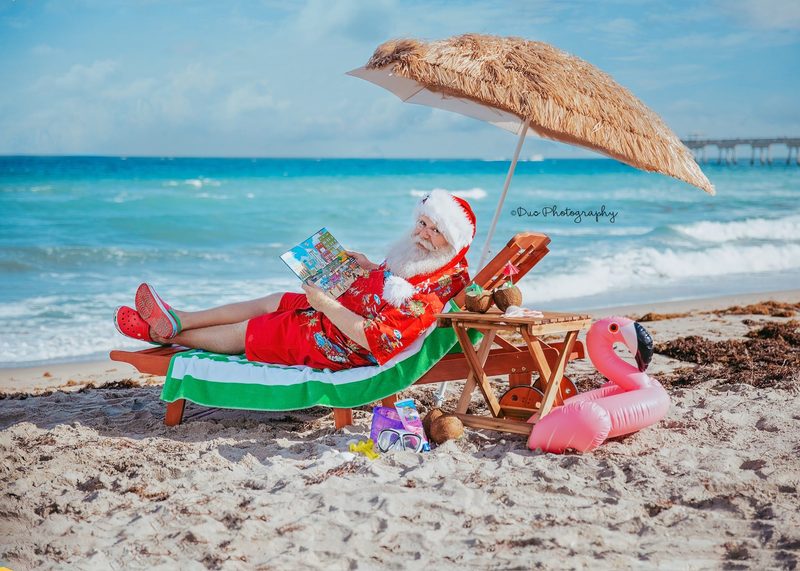 Santa Claus reclining in a beach chair under a thatched umbrella.