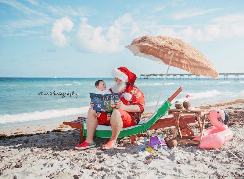 Santa Claus reading on the beach beside turquoise water.