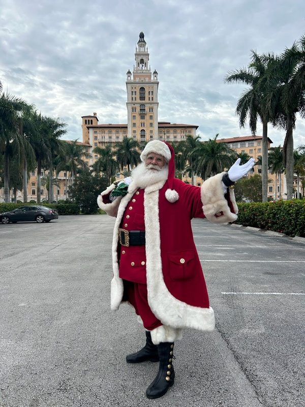 Santa Claus standing before the Biltmore tower in Coral Gables, palm trees behind him.