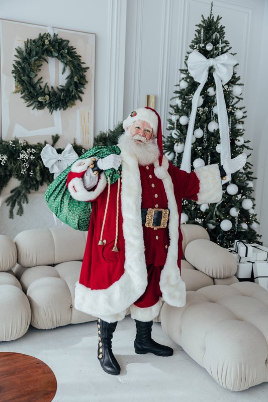 Santa Claus standing head to toe in full red velvet with a white fur trim, a green sack over one shoulder, beside two decorated Christmas trees.
