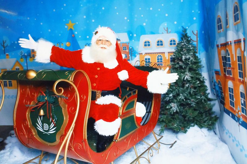 Santa Claus seated in a red and gold sleigh with both arms open wide, against a painted winter village backdrop.
