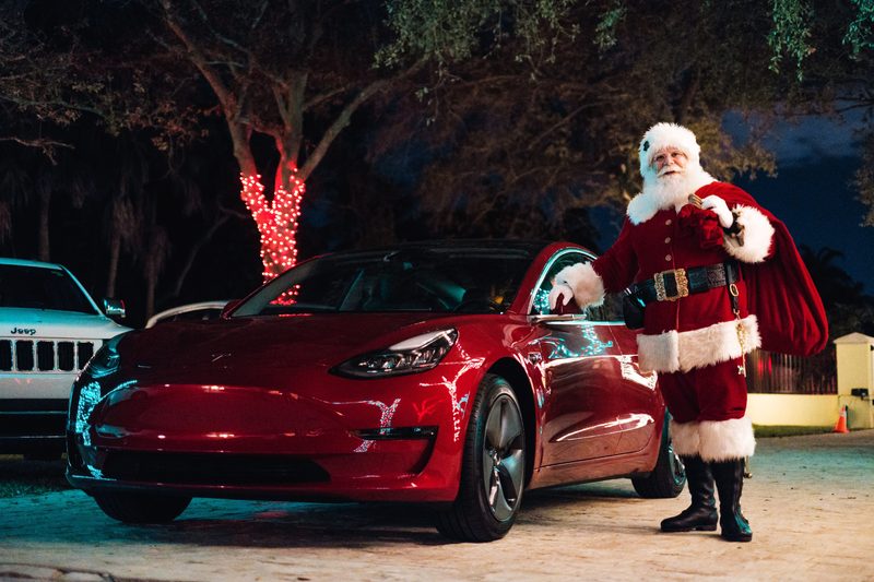 Santa Claus standing beside a red car at night.